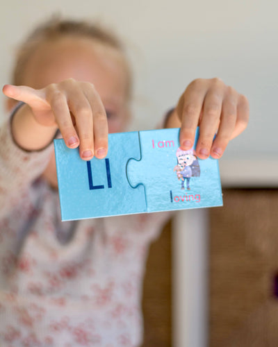 A child holds two connected blue puzzle pieces from the Mindful Champs 'A-Z I Love Me' Affirmation Puzzle, showing “LI,” the phrase “I am loving,” and a cartoon character hugging a heart to inspire positive self-image.