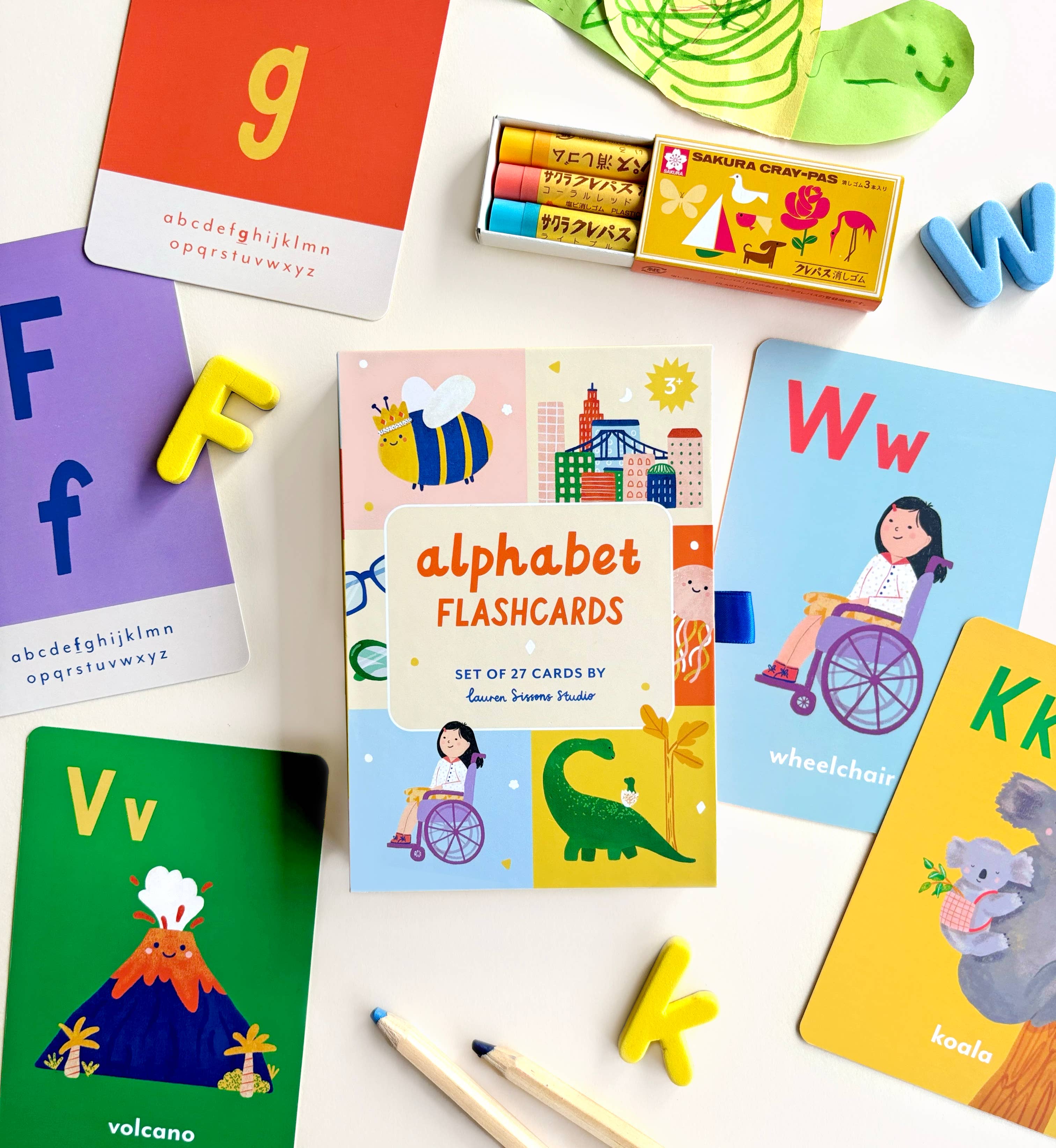 Lauren Sissons Studio's A-Z Illustrated Alphabet Flashcards are displayed with crayons and pencils on a white surface. Each card features large letters, fun illustrations, and words like volcano, wheelchair, and koala for kids’ learning.