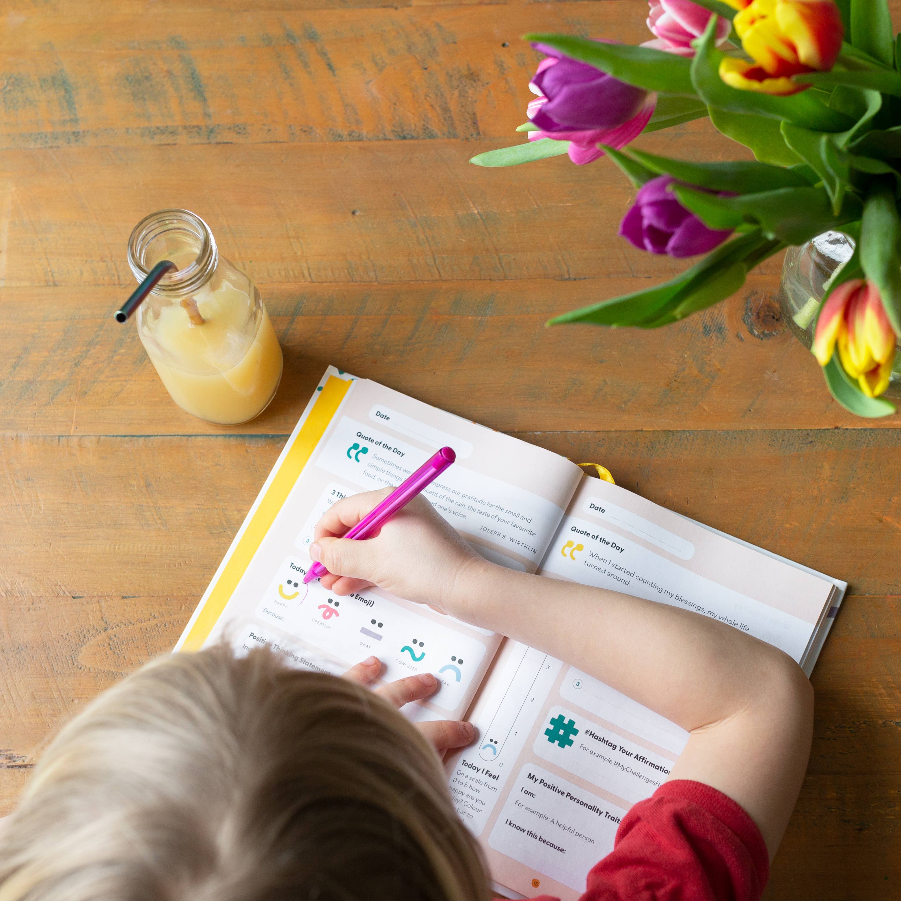 A blonde child writes in the My Mindful Me Journal by Mindful Champs with a pink pen at a wooden table. Beside the open journal are a glass bottle of juice with a straw and a vase filled with colorful tulips.
