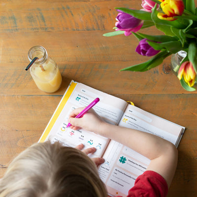 A blonde child writes in the My Mindful Me Journal by Mindful Champs with a pink pen at a wooden table. Beside the open journal are a glass bottle of juice with a straw and a vase filled with colorful tulips.