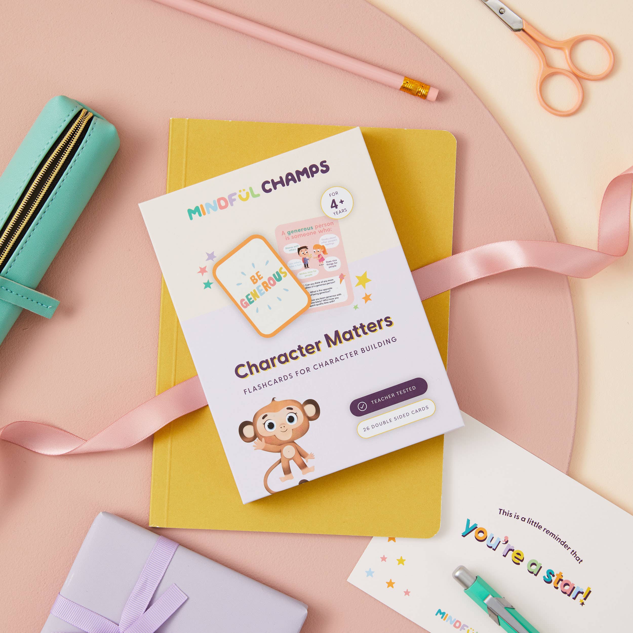 A flat lay shows Mindful Champs’ Character Matters Flashcards for Kids on a yellow notebook with a mint pencil case, pencil, pink ribbon, purple gift box, and a “you’re a star!” card among colorful stationery for personal growth.