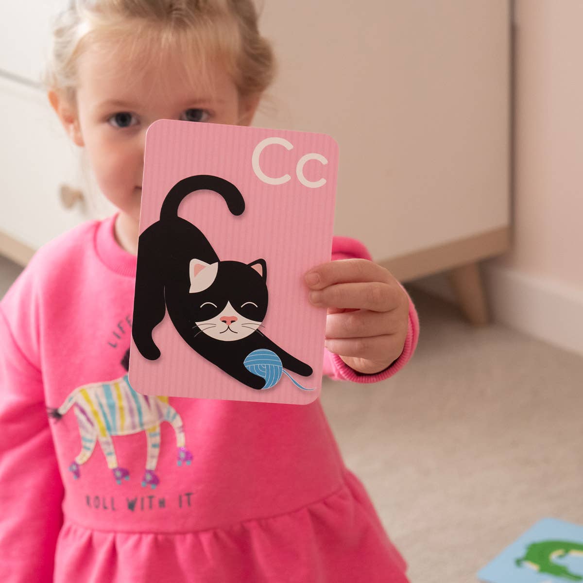 A young child in a pink dress holds up a Priya & Peanut Alphabet Flash Card featuring the letter C and an illustration of a black and white cat with a blue ball of yarn from the "Learn The Alphabet Easily" set.