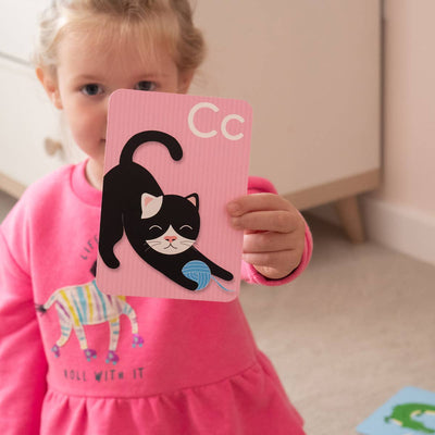 A young child in a pink dress holds up a Priya & Peanut Alphabet Flash Card featuring the letter C and an illustration of a black and white cat with a blue ball of yarn from the "Learn The Alphabet Easily" set.