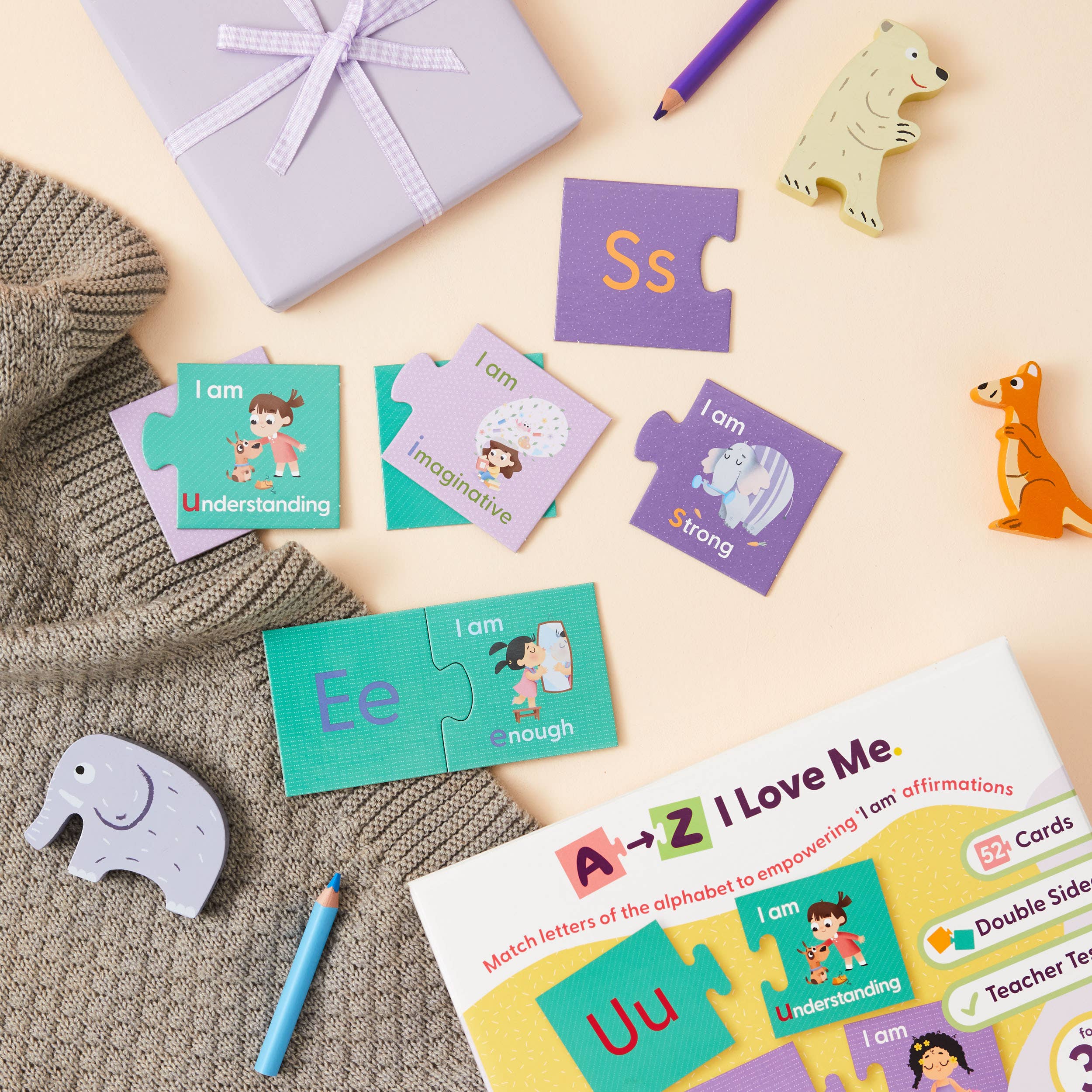A flat lay featuring Mindful Champs’ 'A-Z I Love Me' Affirmation Puzzle, a wrapped gift, animal figurines, sweater, and pencils. This alphabet matching game builds kids’ confidence with affirmations like “I am Understanding” and “I am Strong.”.