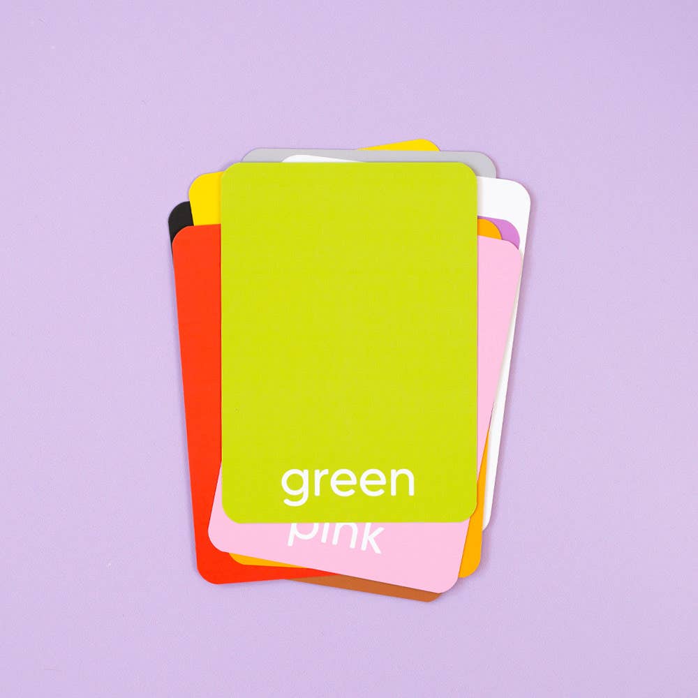 A stack of Priya & Peanut Colour Flash Cards with rounded corners is displayed on a light purple surface. Ideal for early learning, the top card reads "green," while red, orange, pink, white, and yellow cards are also visible.