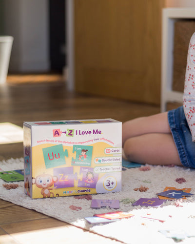A child sits on a rug by the Mindful Champs 'A-Z I Love Me' Affirmation Puzzle box, an alphabet matching game with affirmation cards in playful designs, building positive self-image as cards are spread out under sunlight.