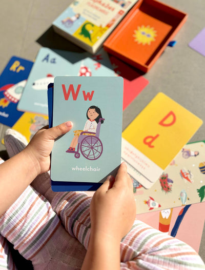 A child explores Lauren Sissons Studio’s A-Z Illustrated Alphabet Flashcards, holding the Ww for wheelchair card featuring a girl in a wheelchair. Other vibrant cards and books create a fun, engaging learning space for kids.