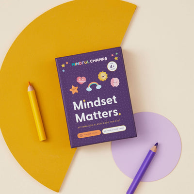 A purple Mindset Matters Flashcards box by Mindful Champs, designed to boost kids' self-esteem and promote a growth mindset, sits on a pastel yellow and purple background with yellow and purple pencils nearby.
