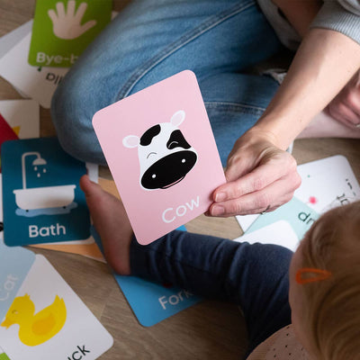 An adult and child sit with Priya & Peanut’s First Word Flash Cards | 50 First Words For Toddlers; the adult holds a pink card with a cartoon cow and “Cow,” helping support toddler speech development through colorful, playful learning.
