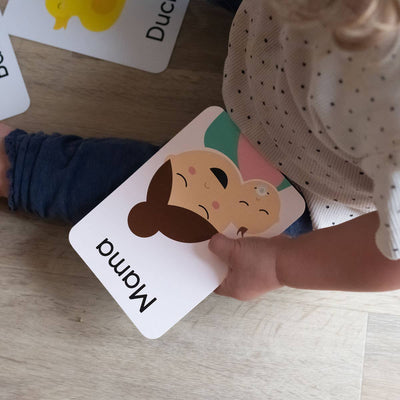 A young child holds a Priya & Peanut First Word Flash Card labeled Mama, while other cards from the 50 First Words For Toddlers set, such as a yellow duck, are scattered nearby to support speech development and playful learning.