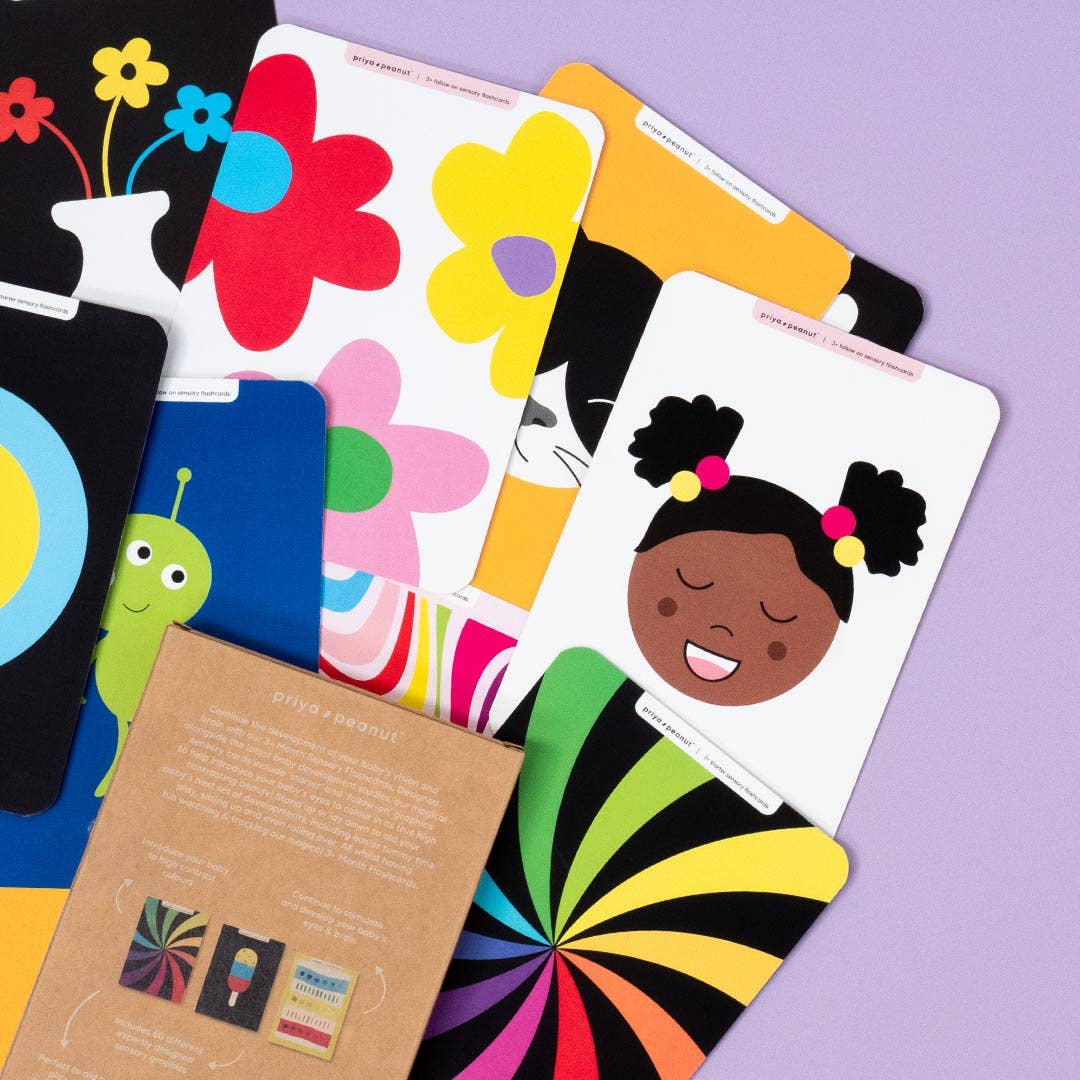 Priya & Peanut’s Colour Sensory Flash Cards For Babies (3+ Months) are fanned out on a purple surface, showing vibrant flowers, a smiling girl with pigtails, a green alien, and bold patterns next to brown packaging.