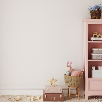 A cozy children’s room features a pink shelf, wicker basket with bunny doll, pink suitcases, wooden blocks, star decor, and a plant. The look is completed with Benji & Belle’s Striped Half Wall Bespoke Wallpaper Panels on the white wall.