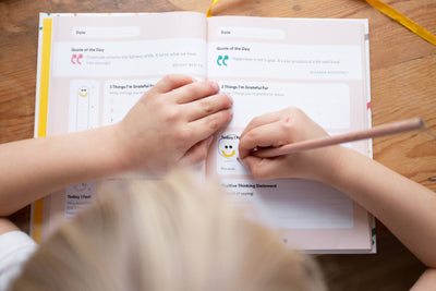 A child writes with a pencil in the My Mindful Me Journal by Mindful Champs, filling out gratitude prompts at a wooden table as another child's hand rests gently on the open page.