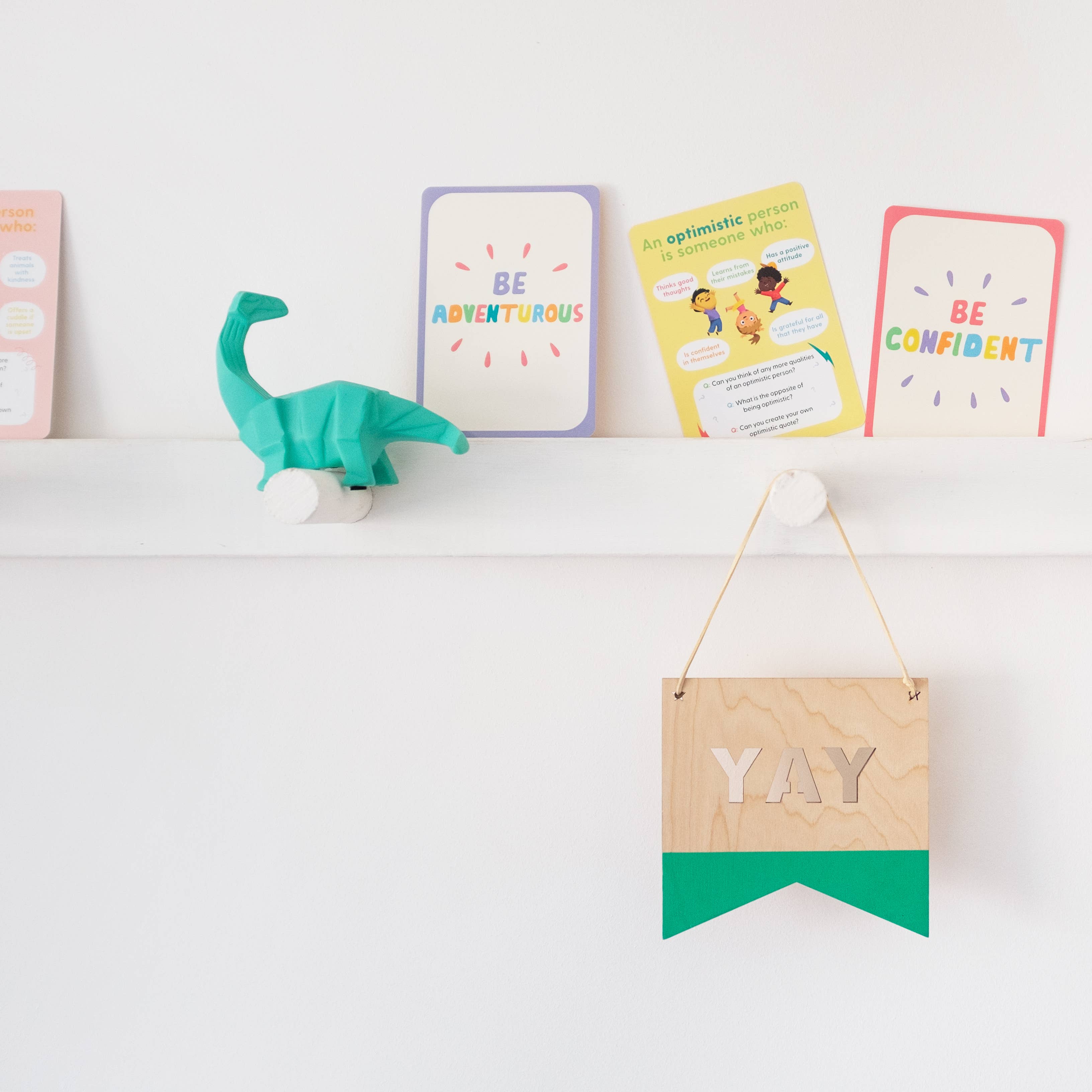 A white shelf holds a green dinosaur figurine, a set of Mindful Champs Character Matters Flashcards for Kids, and a wooden sign with a green bottom edge reading "YAY" hanging below. Both the shelf and wall are white.