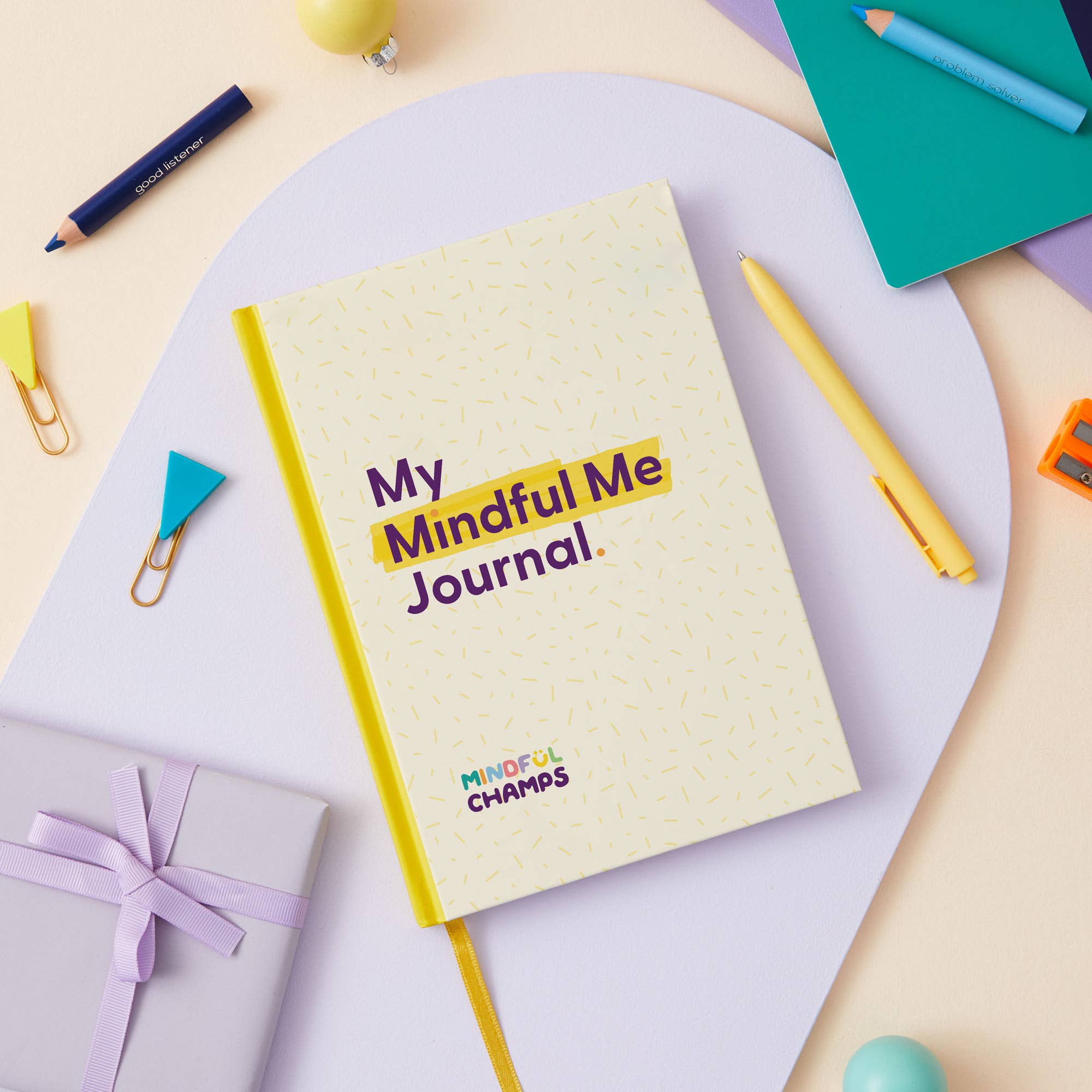 The My Mindful Me Journal by Mindful Champs, a yellow mindfulness journal for kids with dotted patterns on the cover, lies on a white surface surrounded by colorful stationery and a wrapped gift—ideal for supporting children's mental health.