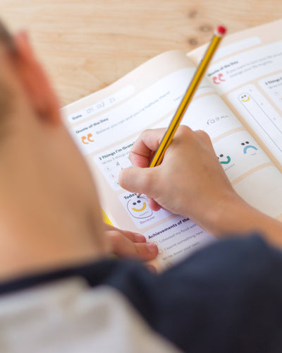 A child uses a yellow pencil to fill out prompts and draw in the open "My Mindful Me Journal" by Mindful Champs, focusing on gratitude and mindfulness practices.