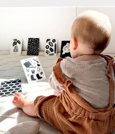 A baby in a beige shirt and rust romper sits on a soft surface, facing away, looking at Lauren Sissons Studio’s High Contrast Flashcards for Baby—Sensory Newborn Gift—displayed against a wall in bright natural light.