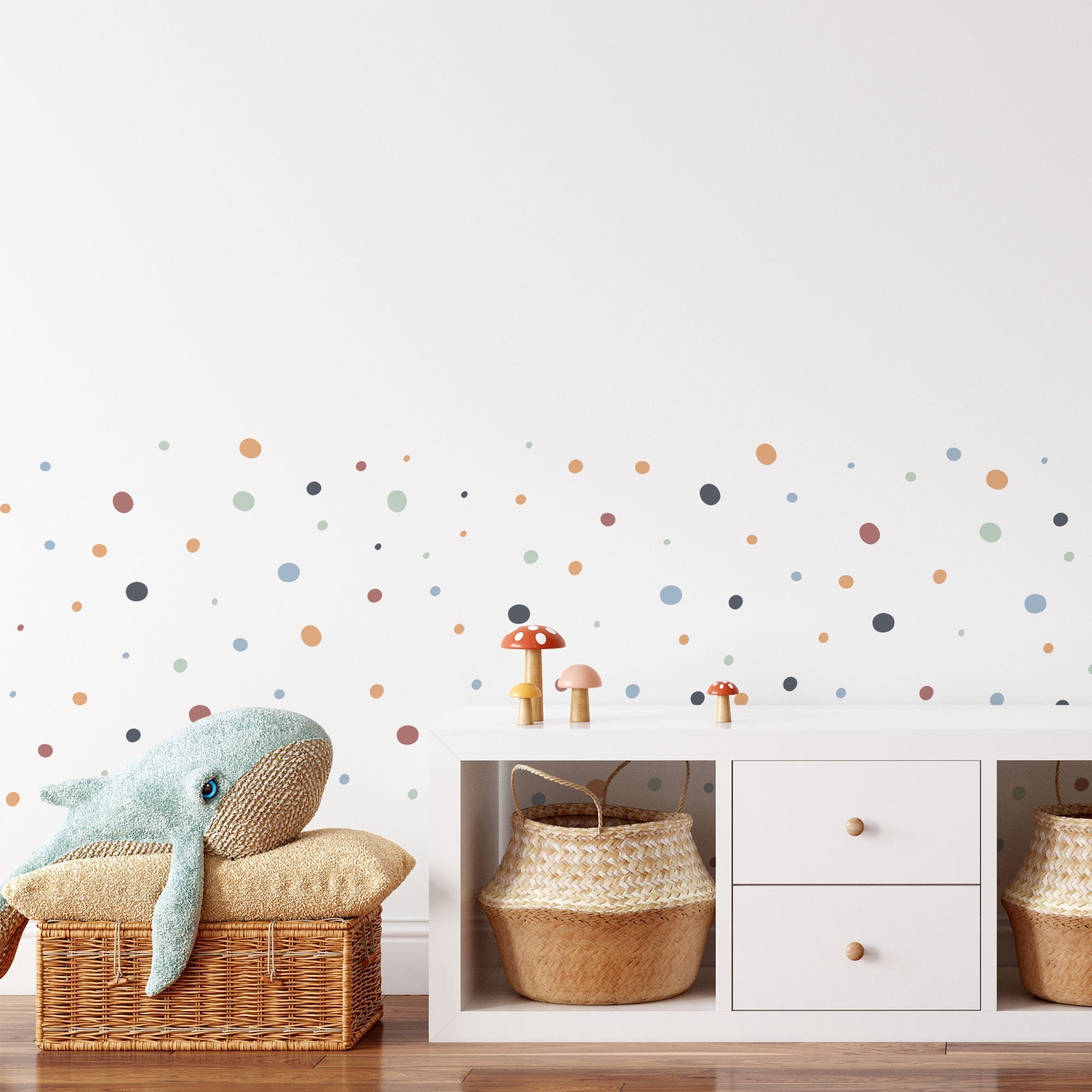 A minimalist childrens room has a white wall with My Stores Boho Polka Dots Stickers. A wicker basket holding a plush whale is next to a white cabinet with two woven baskets and whimsical mushroom decorations on top.