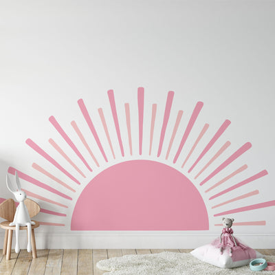 The kids room has a large Boho Sun Sticker by My Store on the wall. A plush rabbit is on a white chair to the left, and a teddy bear is on a pink pillow over a fluffy white rug. Warm wooden flooring finishes the nursery decor.