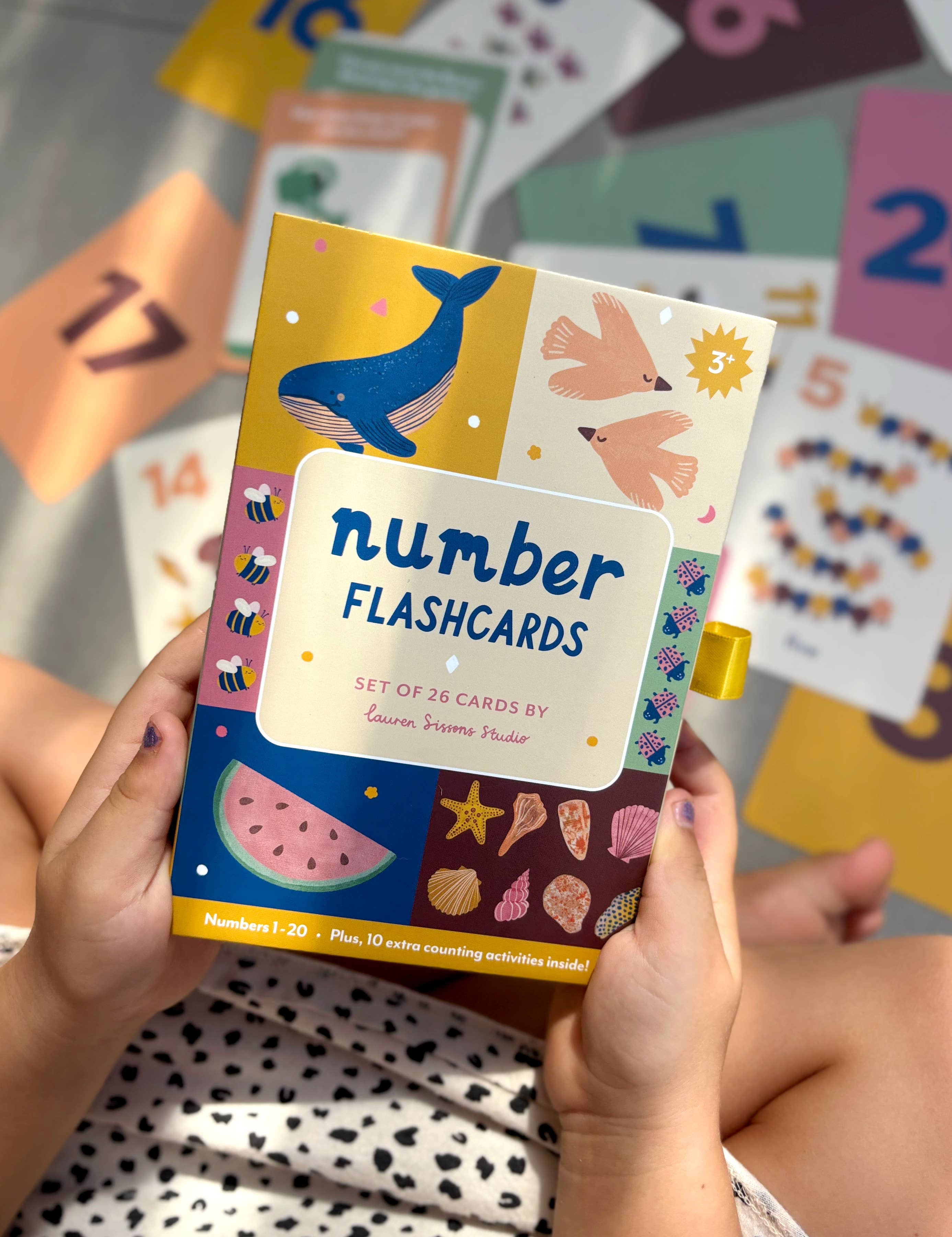 A child holds a colorful box of Lauren Sissons Studio's Illustrated Number Flashcards - Kids Toys Learning Education, featuring playful illustrations like whales and birds. Number cards (1, 10, 17, 21, etc.) are scattered in the background.