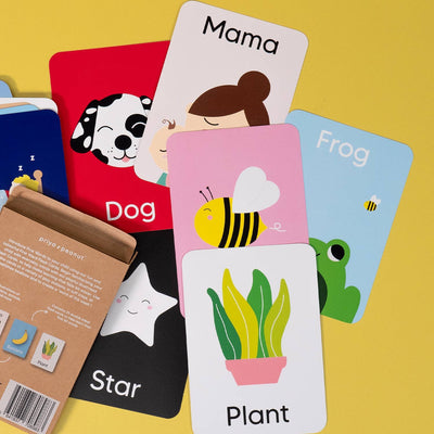 Priya & Peanut's First Word Flash Cards | 50 First Words For Toddlers are displayed on a yellow surface featuring words and drawings like dog, Mama, bee, frog, star, and plant—ideal for supporting toddler speech development. Box with instructions shown.