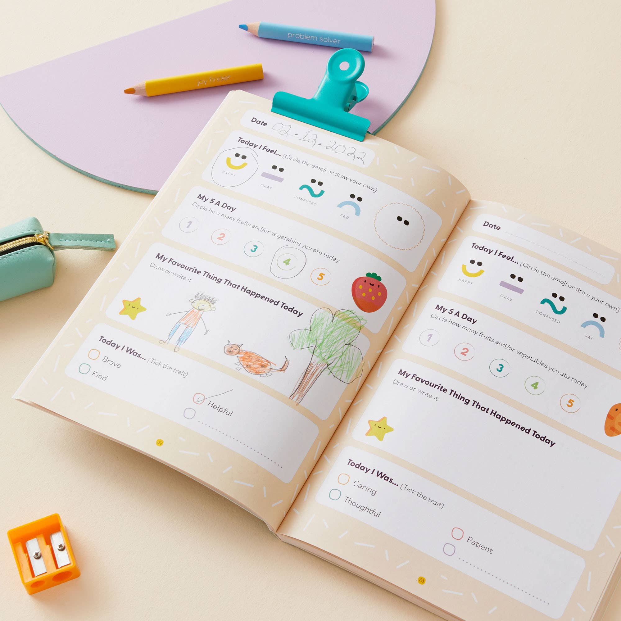 The My Mindful Me Journal Early Years Edition by Mindful Champs is an open feelings journal for kids with prompts for moods, favorites, and emotions. Sun, flower, and fruit drawings decorate the pages with colored pencils and accessories nearby.