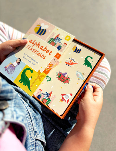 A child holds a bright box of Lauren Sissons Studio’s A-Z Illustrated Alphabet Flashcards—kids’ toys decorated with playful animal, object, and building images for fun learning. The child wears striped leggings and a denim jacket.
