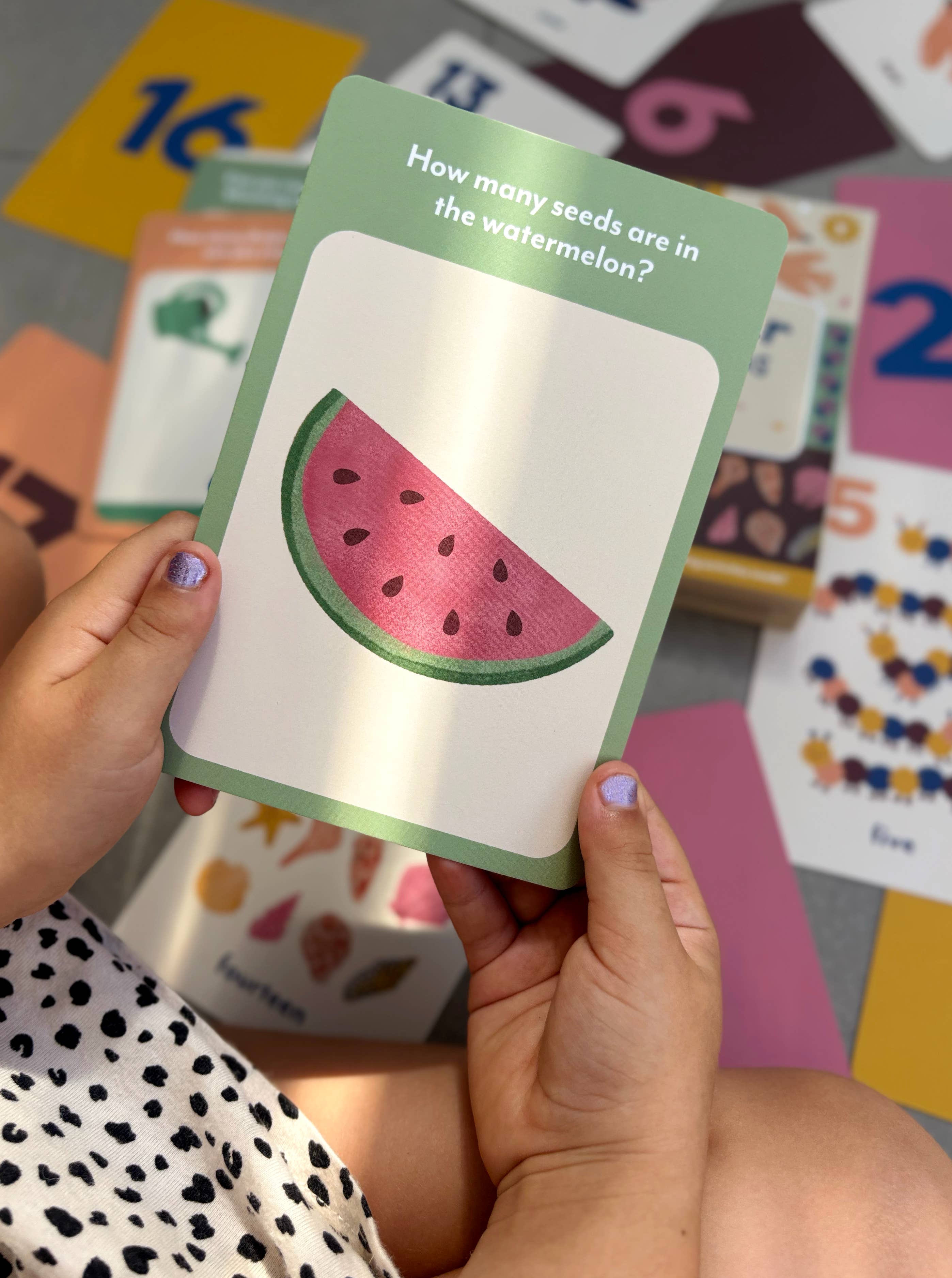 A child holds an Illustrated Number Flashcard by Lauren Sissons Studio featuring a watermelon slice and “How many seeds are in the watermelon?” Colorful number flashcards from this learning set are scattered on the floor.