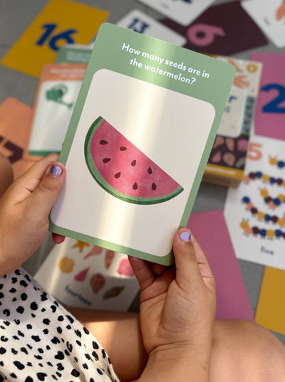 A child holds an Illustrated Number Flashcard by Lauren Sissons Studio featuring a watermelon slice and “How many seeds are in the watermelon?” Colorful number flashcards from this learning set are scattered on the floor.