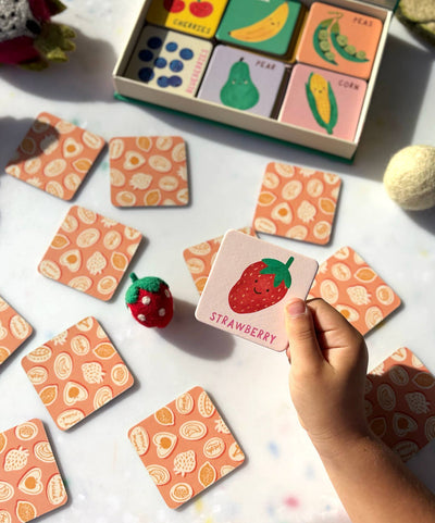 A child holds a card with a strawberry illustration and the word “Strawberry” from the Fruit and Veg Memory & Matching Game by Lauren Sissons Studio, as other matching fruit cards invite fun educational play on the table.