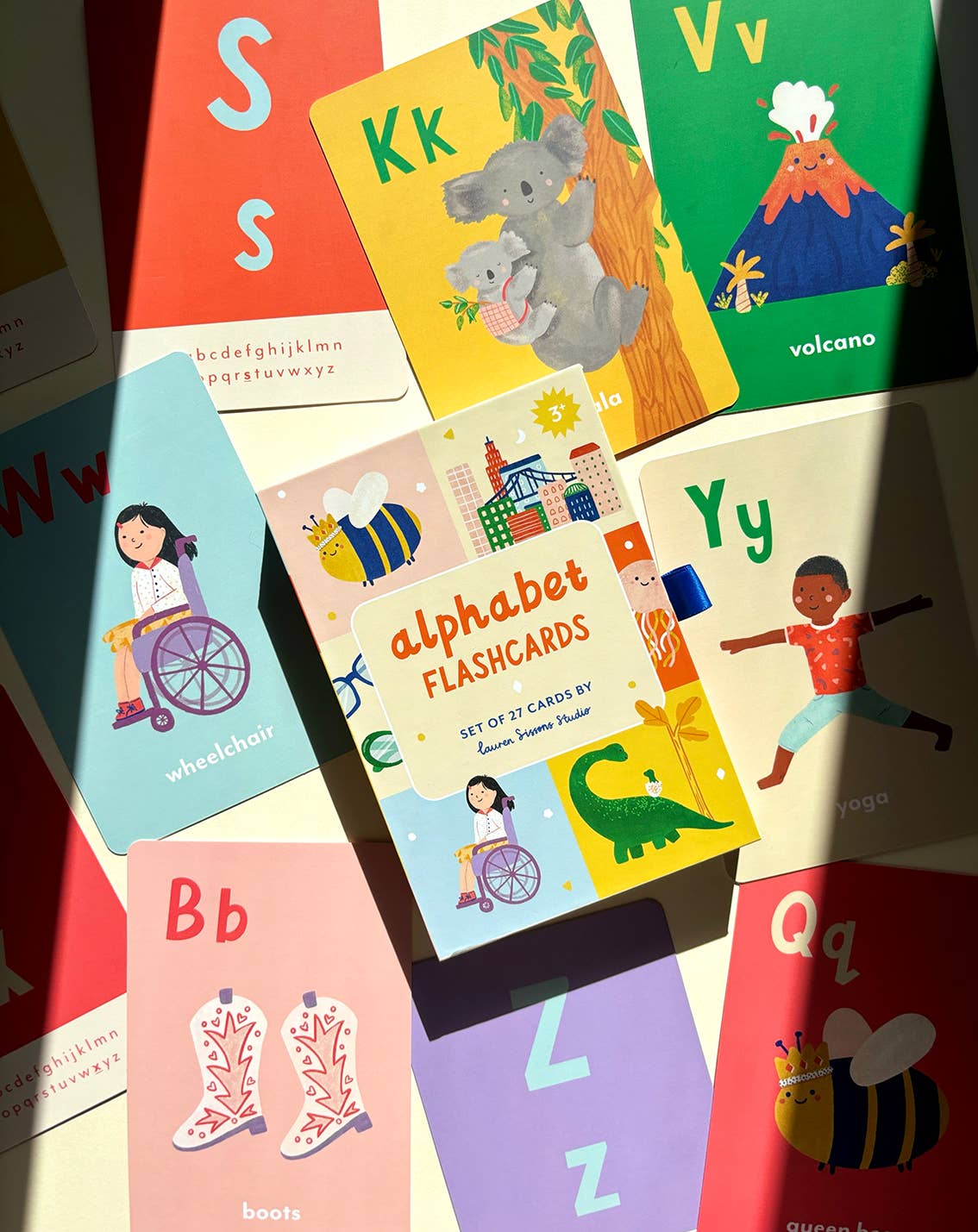 A-Z Illustrated Alphabet Flashcards by Lauren Sissons Studio are spread out with playful images like a volcano, wheelchair, yoga pose, boots, and koala. The central box labeled "alphabet FLASHCARDS" makes learning fun for kids.
