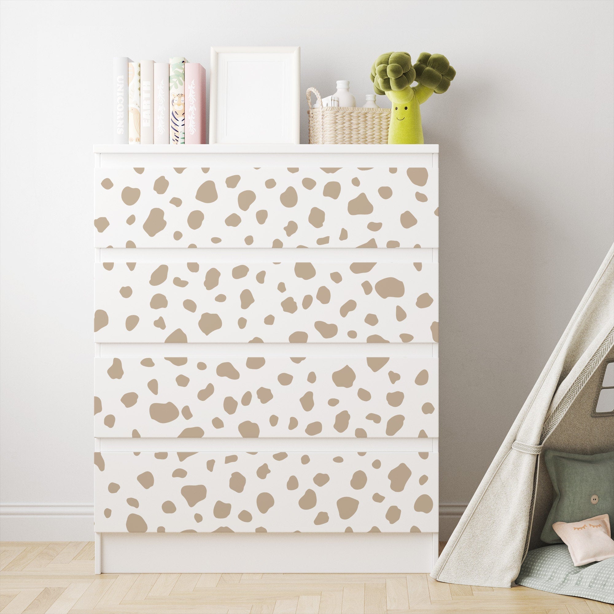 A white dresser, accented with My Stores Dalmatian Spot Stickers, stands against a light wall. Topped with books, a woven basket, and a green plush toy, it exudes charm. Nearby, a gray tent sits on the wooden floor, adding to the delightful setup.