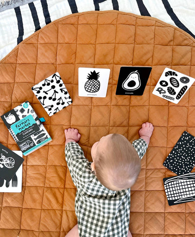 A baby in a checkered outfit lies on a quilted brown mat, exploring Lauren Sissons Studio’s High Contrast Flashcards for Baby—featuring cherry, pineapple, avocado, and bread images—for sensory newborn development. Other cards and the product box are nearby.