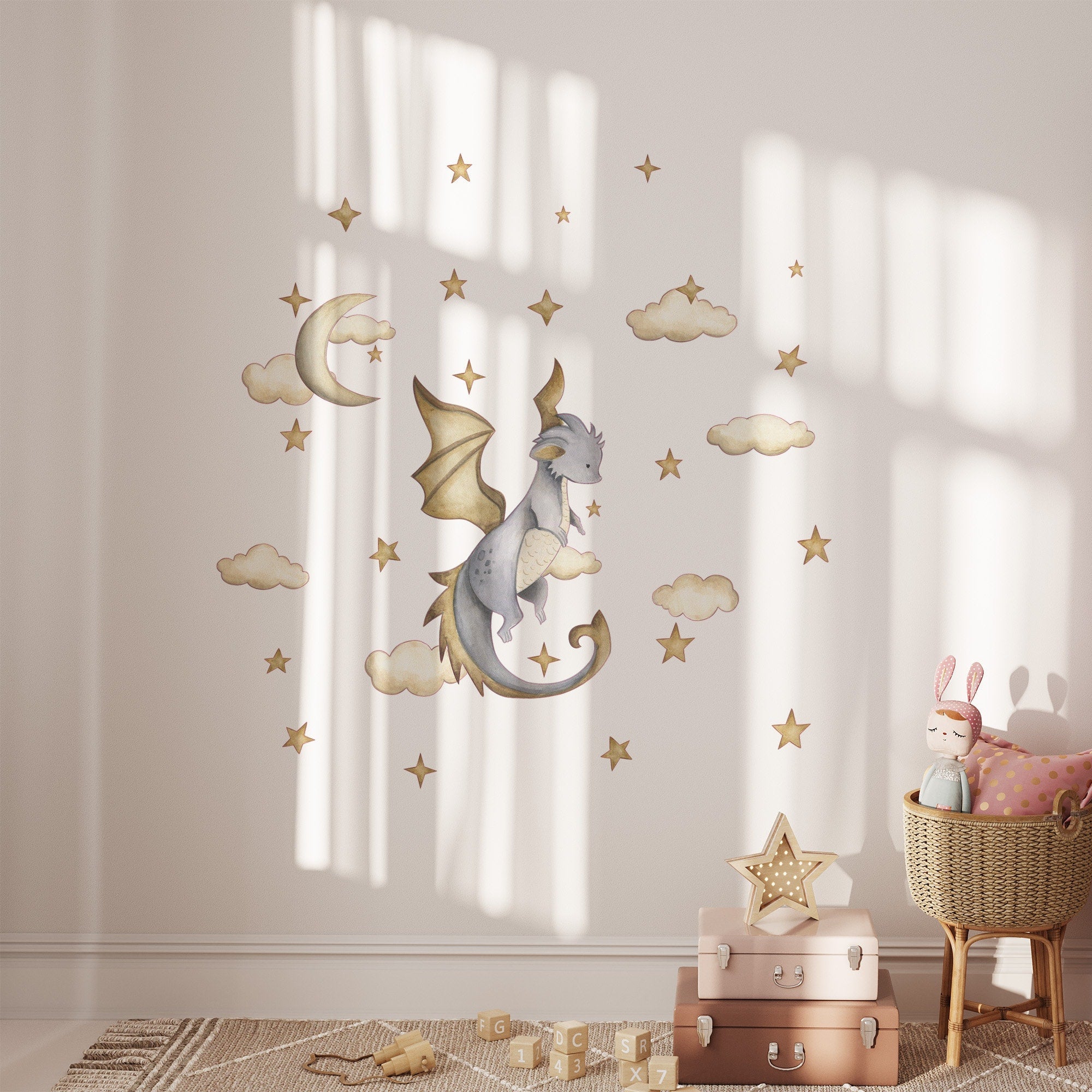 Soft light fills the cozy nursery. My Stores Dragon Stickers, featuring a whimsical dragon, clouds, a crescent moon, and stars grace the wall. A plush bunny rests on pink cushions beside stacked suitcases topped with a star-shaped lamp.