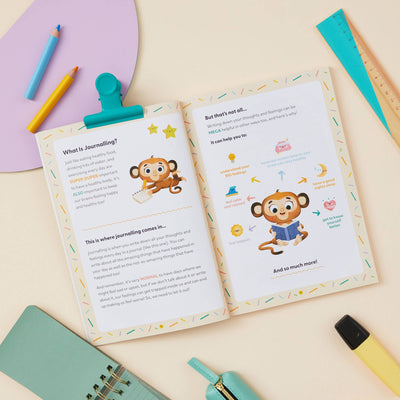 The My Mindful Me Journal Early Years Edition by Mindful Champs lies open on a desk, its playful monkey illustrations and easy text introducing kids to journaling. Colorful pens, a ruler, and a teal notebook surround the book.
