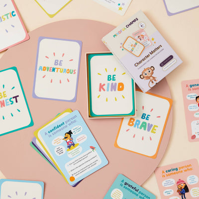 Mindful Champs' Character Matters Flashcards for Kids are displayed on a round pink surface, showing positive messages like Be Adventurous, Be Kind, and Be Brave. The deck's box features a cartoon monkey.