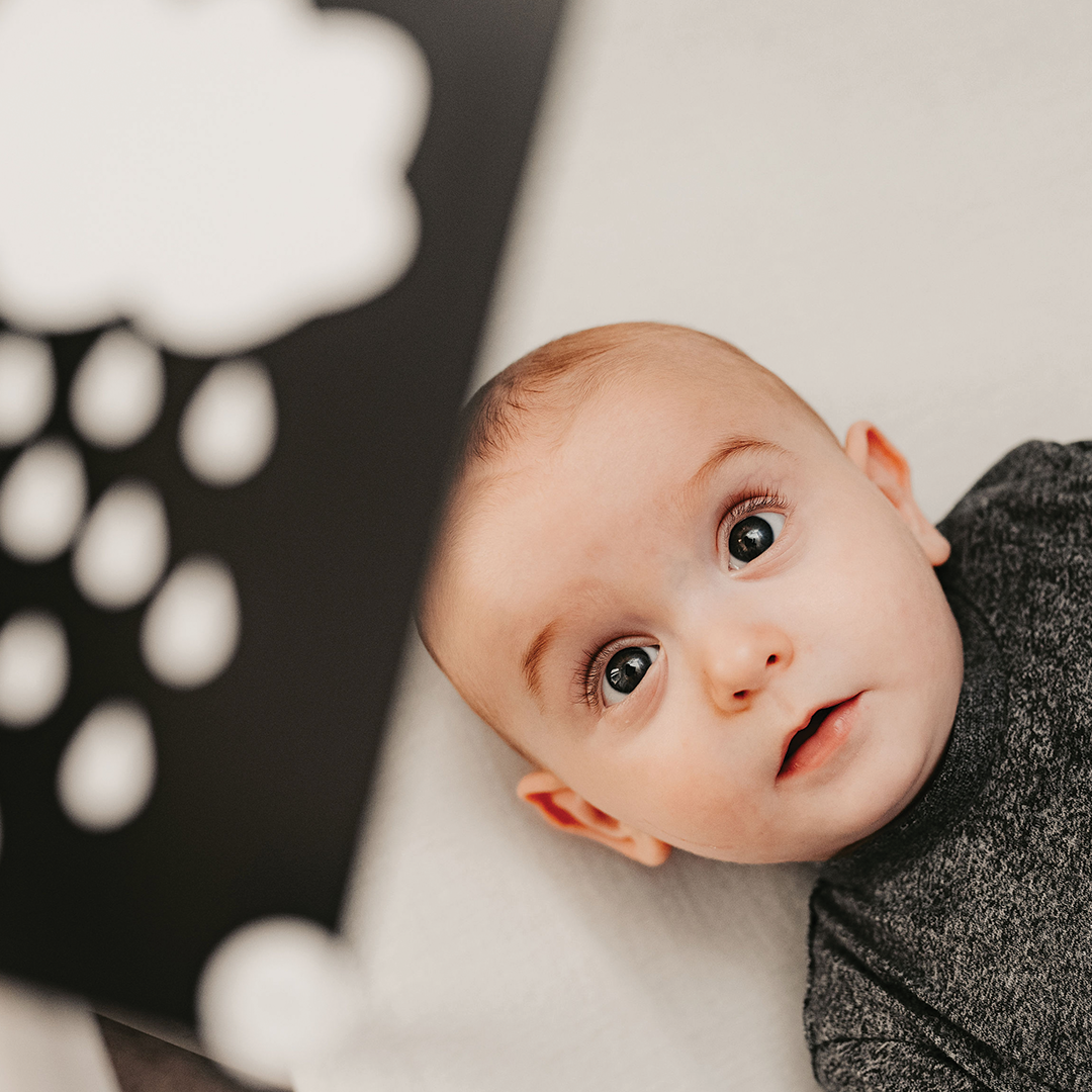 A curious baby in a dark outfit lies on a light surface, gazing up at Priya & Peanut’s Newborn Sensory Cards (0+ Months Old) — an award-winning baby toy designed for early visual development with bold, high-contrast shapes.