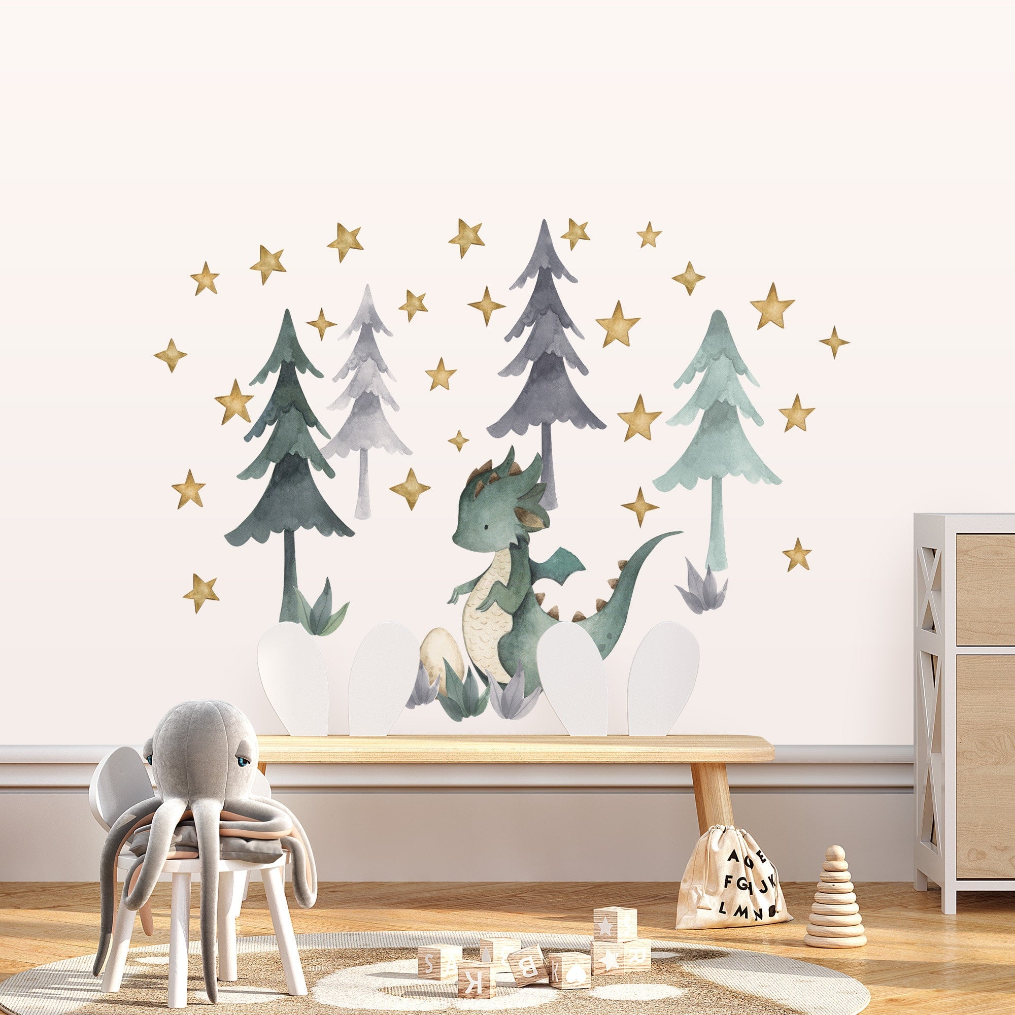 A childrens room decorated with My Stores Green Dragon Stickers on a wall, depicting dragons among pine trees and stars. The space includes a plush octopus, wooden alphabet blocks, and stackable toys on a light rug beside a wooden dresser for easy and fun decoration.