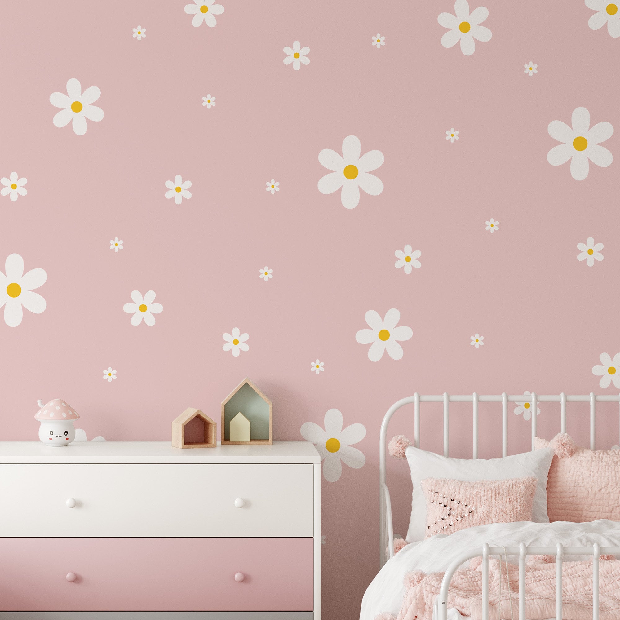 A childs room is decorated with My Stores Daisy Wall Decals and pink-white floral wallpaper. It includes a white metal bed with pink bedding, a pink and white dresser with mushroom ornaments, and small wooden houses on top—a lovely nursery haven.