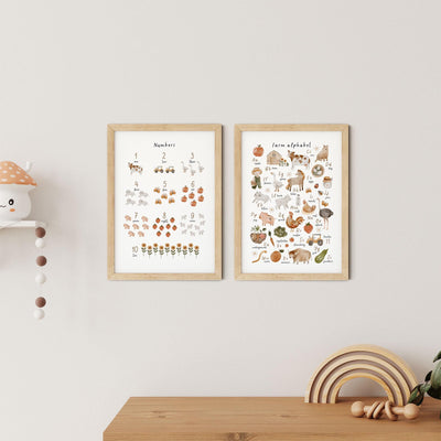 Two framed Farm Animals Alphabet & Numbers Nursery Print by My Store hang on a white wall above a wooden shelf. The left print features numbers with colorful illustrations, while the right showcases a farm-themed alphabet. A small mushroom and wooden rainbow adorn the shelf below.