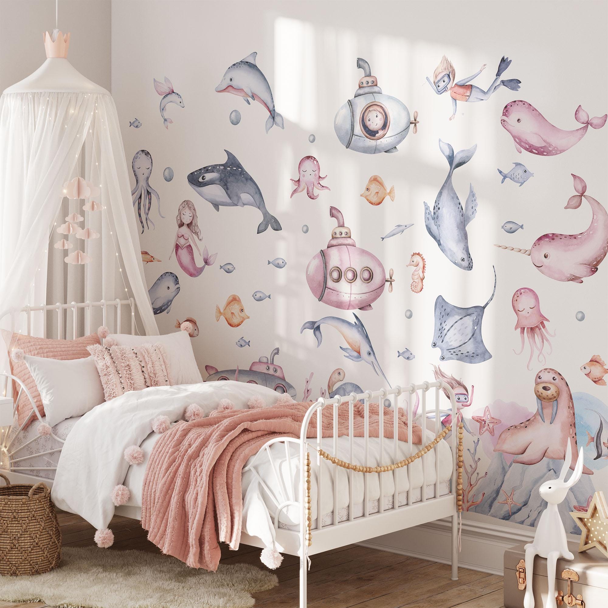 A cozy childs bedroom with a white and pink color scheme features My Stores playful Under The Sea Nursery Wall Stickers, showcasing dolphins, whales, and octopuses. A soft pink blanket and cushions adorn the white bed, with a canopy elegantly draped above.