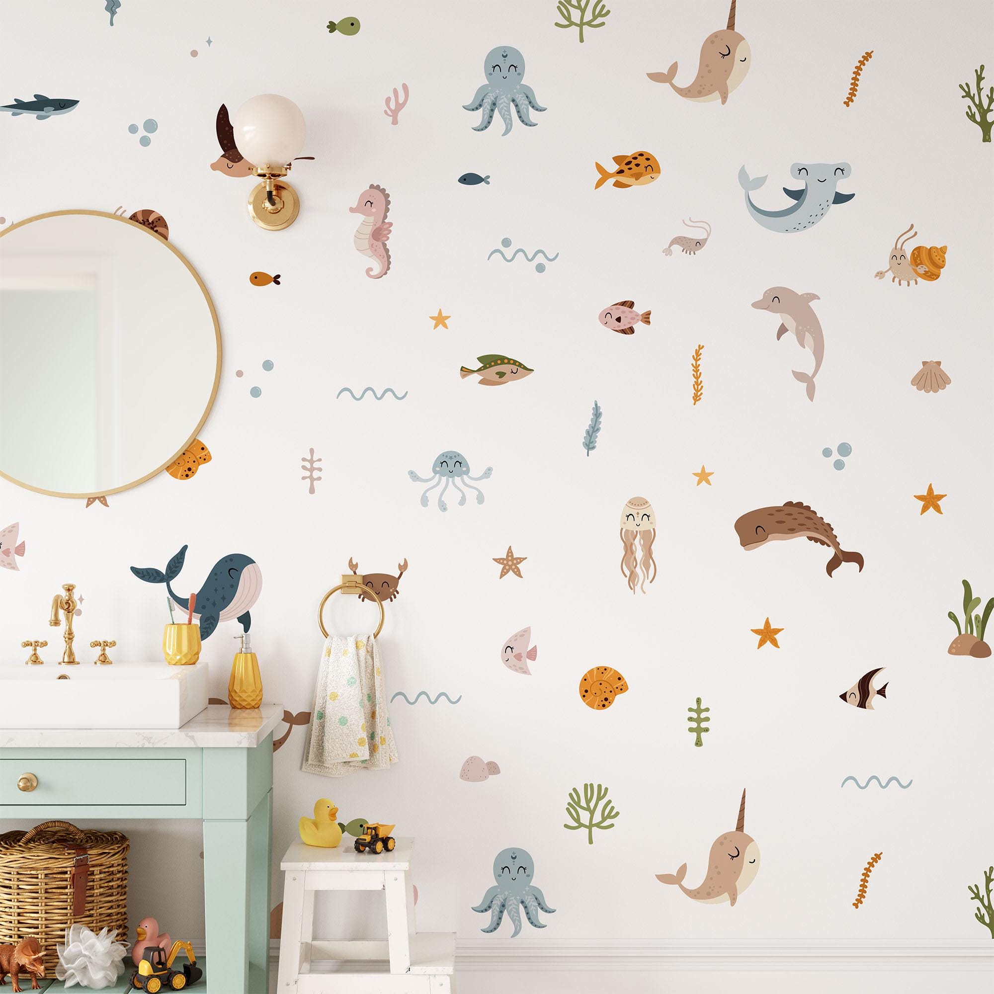 The bathroom wall is decorated with My Stores Ocean Animals Stickers featuring whales, octopuses, seahorses, fish, and seaweed. Above the sink hangs a round mirror beside toiletries under a sleek light fixture. A small cabinet with a basket and toys complements the aquatic theme.
