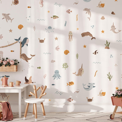 A childrens room with a sea-themed wall design features My Stores Ocean Animals Stickers, including dolphins, narwhals, and fish that adorn the white wall. A chair, small round table, and vase with flowers add to the cozy decor as soft sunlight filters in.