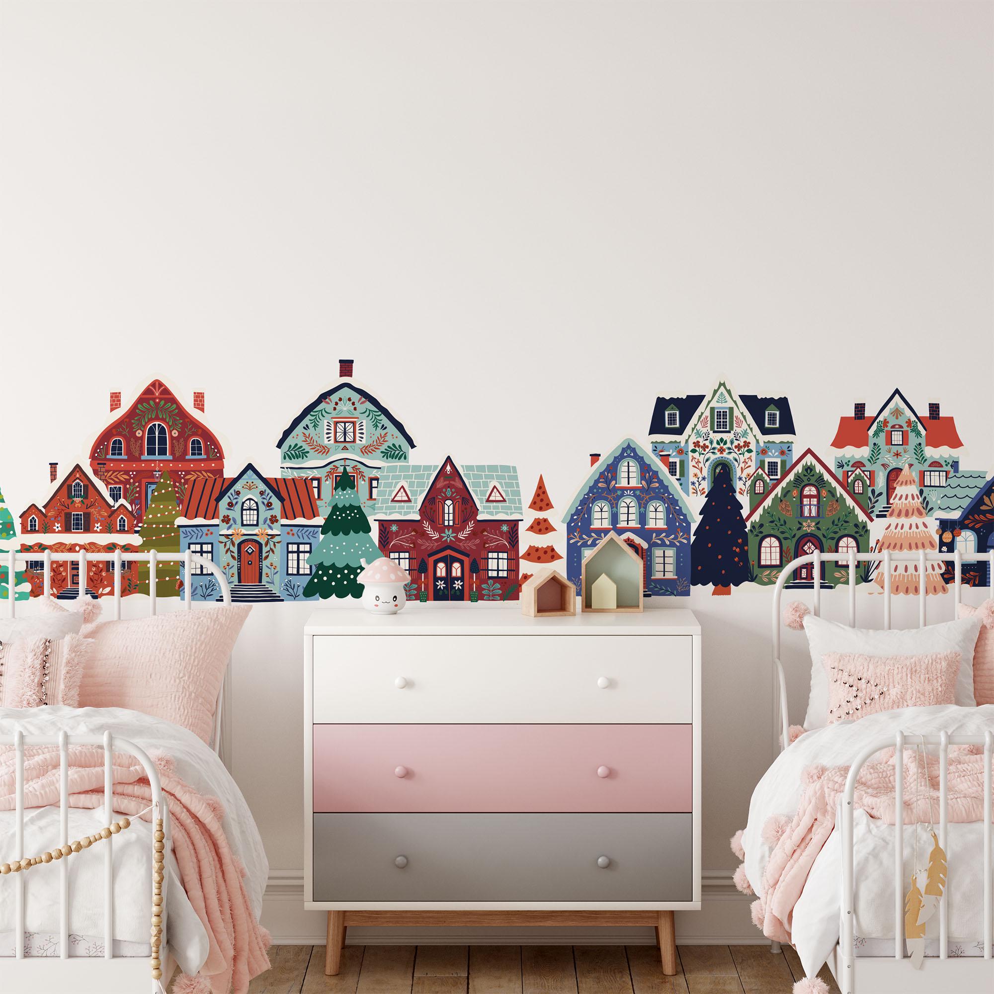 A childrens room has two white metal beds with pink bedding next to a pink and gray dresser. The wall features My Stores Festive Houses Wall Stickers of whimsical cottage-style houses and trees. A small toy bear on the floor adds charm to this festive retreat.