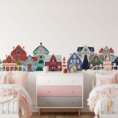 A childrens room has two white metal beds with pink bedding next to a pink and gray dresser. The wall features My Stores Festive Houses Wall Stickers of whimsical cottage-style houses and trees. A small toy bear on the floor adds charm to this festive retreat.
