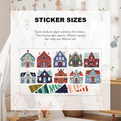 A room with a framed size guide displays vibrant Festive Houses Wall Stickers by My Store, featuring removable colorful houses (~325mm square) and trees (180mm tall), creating a cozy atmosphere with neutral decor.
