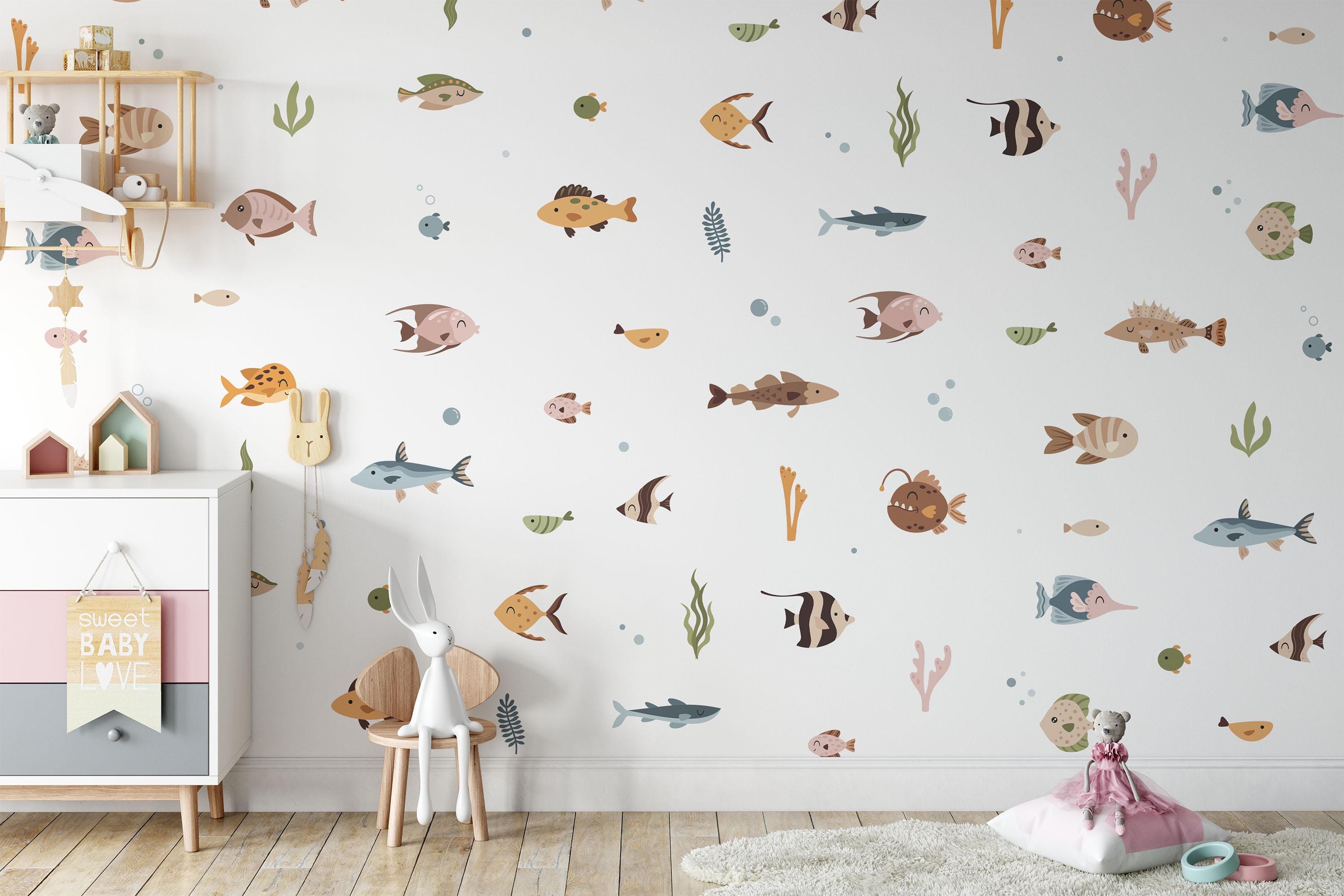 A childrens room has pastel ocean fish stickers from My Store, a white dresser with a lamp and books, a toy rabbit on a wooden chair, and a plush bear on a pink blanket on the floor. Warm wooden flooring completes the cozy ambiance.