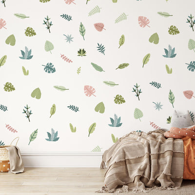 A cozy room with a white wall beautifully decorated using My Stores Tropical Leaves Nursery Wall Stickers in pastel colors. A striped blanket and pillows lay on the floor next to a wicker basket on light wooden flooring, creating a warm and inviting atmosphere.