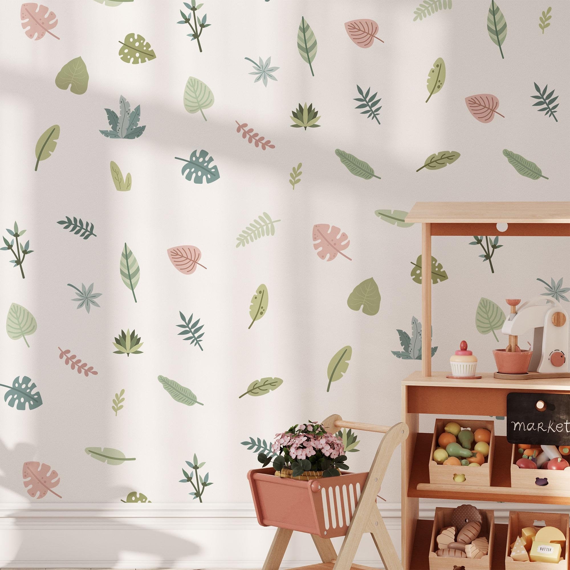 A childrens play area features a market stand with toy fruits and vegetables, enhanced by My Stores Tropical Leaves Nursery Wall Stickers. A basket of pink flowers sits in the foreground, while removable green and brown leaf decals offer easy decor updates.