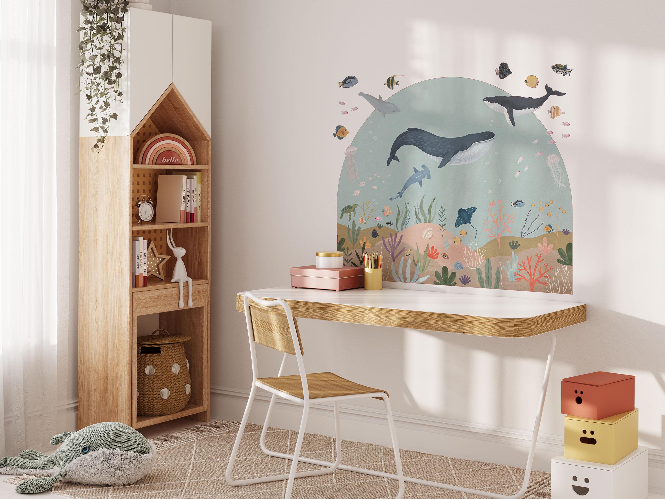 A cozy childrens room with a Sea Animals Arch Wall Sticker by My Store, showcasing whales, fish, and coral. Nearby, a wooden desk and chair sit beside colorful toys and books on shelves. A whale-shaped plush lies on the floor.
